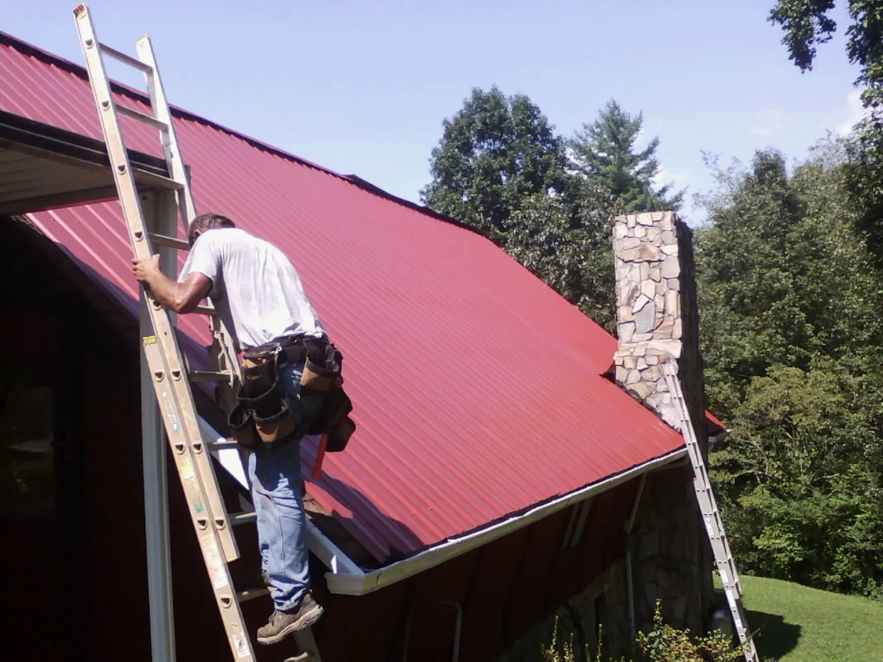 Roofer on a ladder beside a red metal roof and stone chimney — Soffit & Fascia Repair by Roofing Brothers in Catharpin